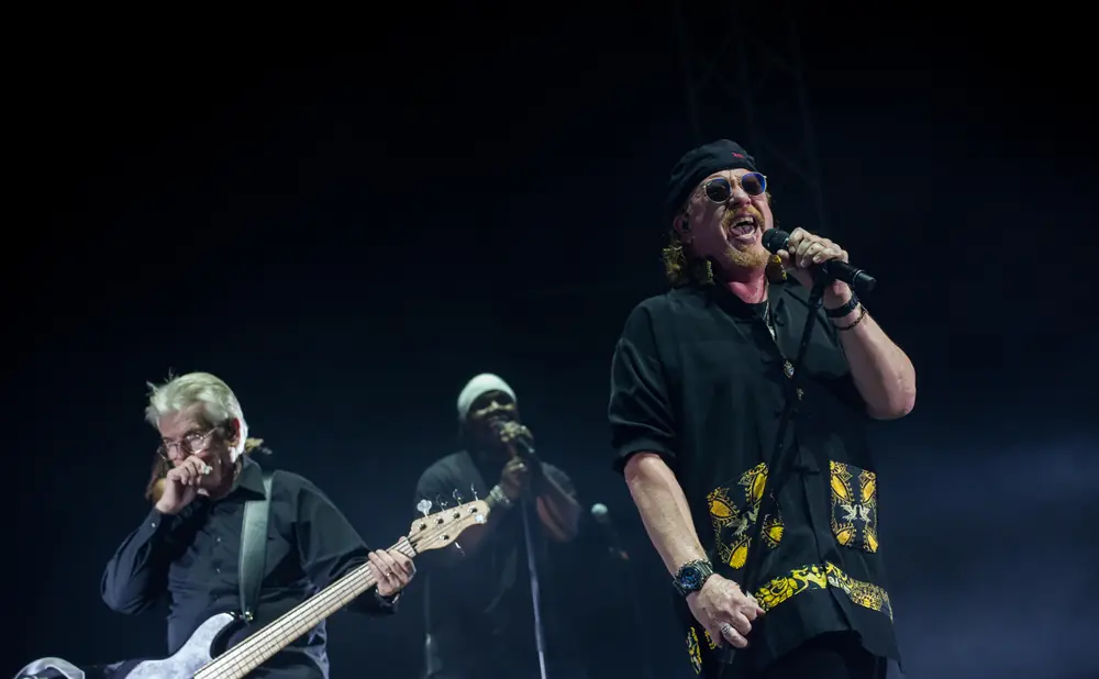 Singer with a red mustache and sunglasses performs onstage with a bassist and backup singer.