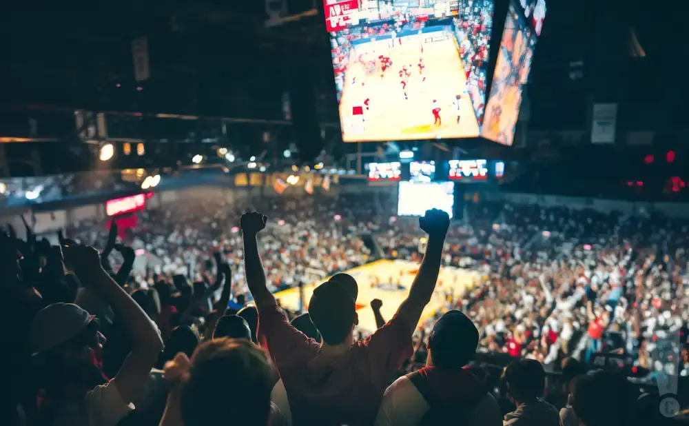 Fans cheer at a basketball game, with a large screen showing the court.