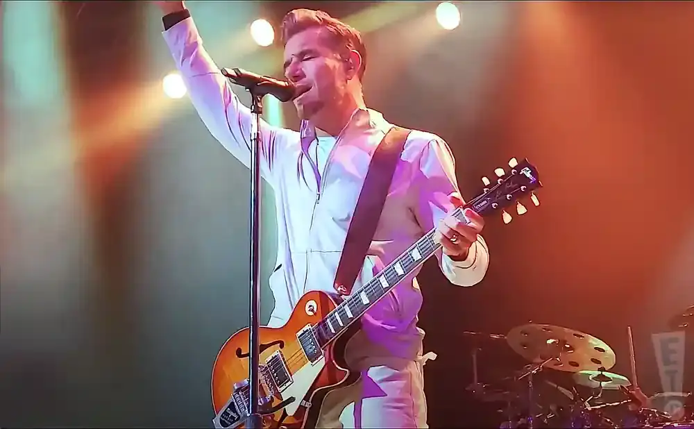 A man in a light-colored tracksuit plays a Gibson Les Paul guitar and sings into a microphone on a stage.