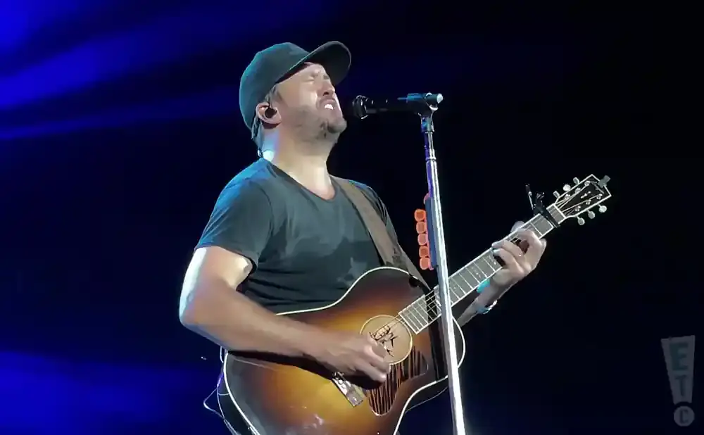 A man in a black t-shirt and baseball cap plays an acoustic guitar and sings into a microphone on a dimly lit stage.