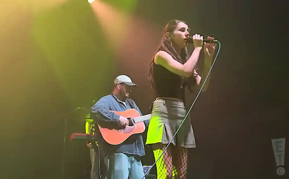 A female singer holds a microphone while a male guitarist plays an acoustic guitar.