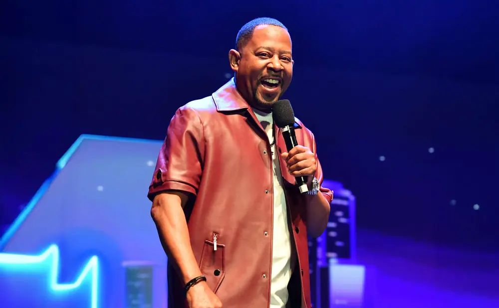 A Black man in a red shirt and pants smiles while holding a microphone on a blue-lit stage.