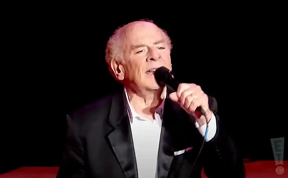 An older man in a suit and white shirt sings into a microphone on a dark stage.