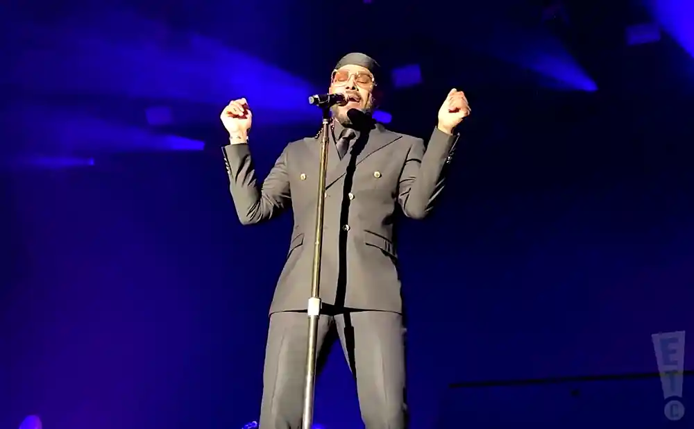 A man in a black suit and head covering sings into a microphone on a blue-lit stage.