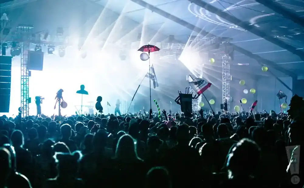 A concert crowd under bright stage lights with balloons and flags in the air.