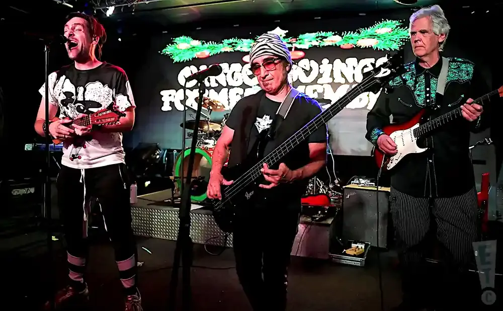 A band plays on stage, with a singer holding a ukulele, a bassist, and a guitarist.