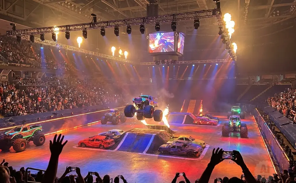 Monster truck show with trucks jumping over cars, flames, and a large crowd watching.