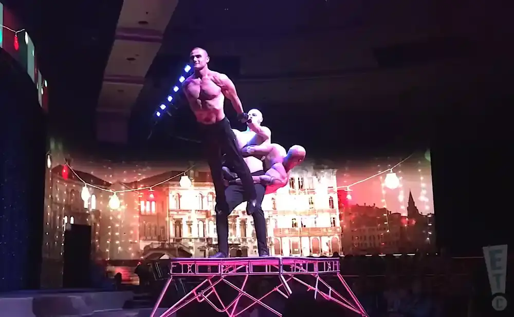 Three muscular men perform a balancing act on a stage, with a painted cityscape backdrop.