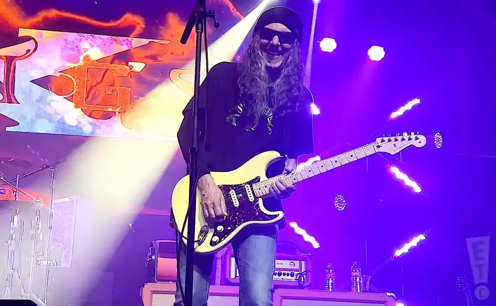 A guitarist with long hair and sunglasses plays a cream-colored Fender Stratocaster on a stage lit by purple lights.