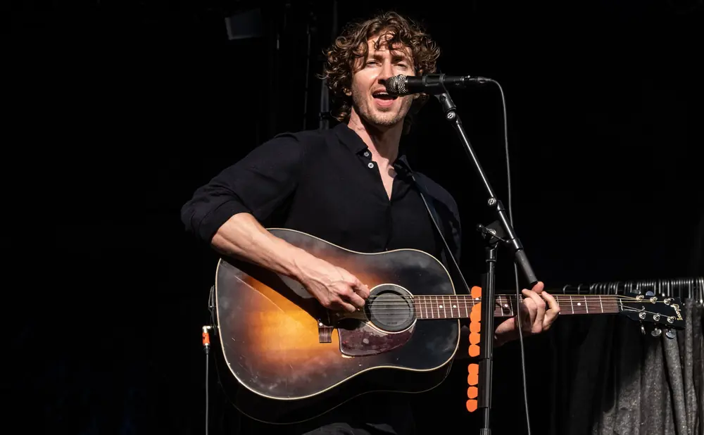 Musician with curly hair sings into a microphone while playing an acoustic guitar.