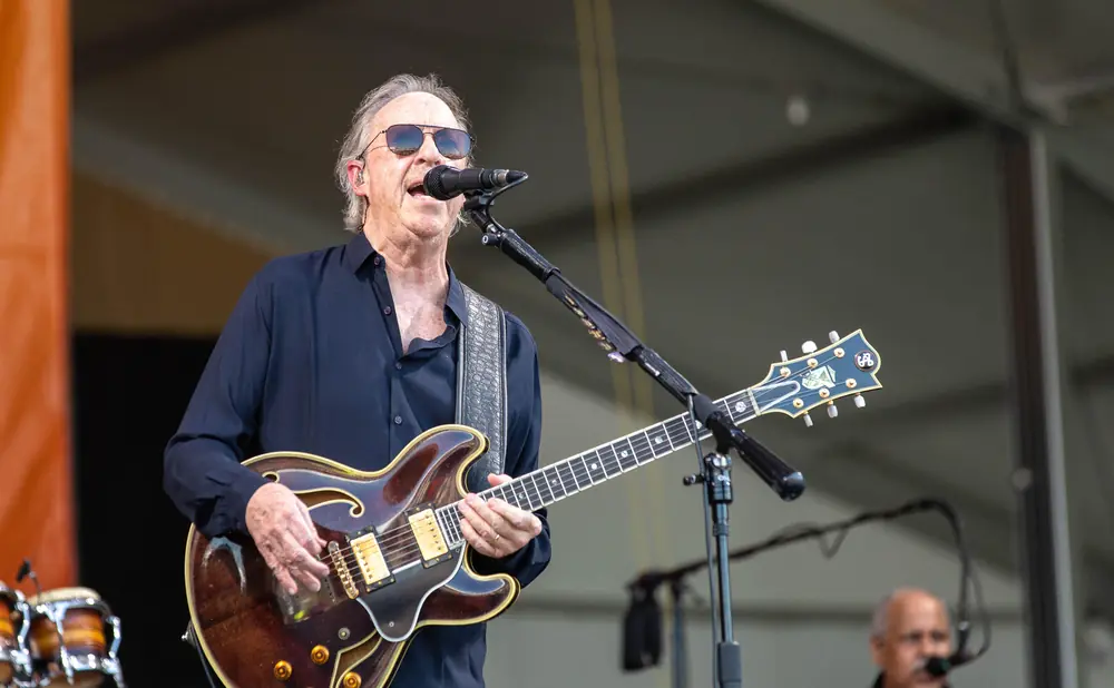 A man in sunglasses plays a brown hollow-body guitar on stage while singing into a microphone.