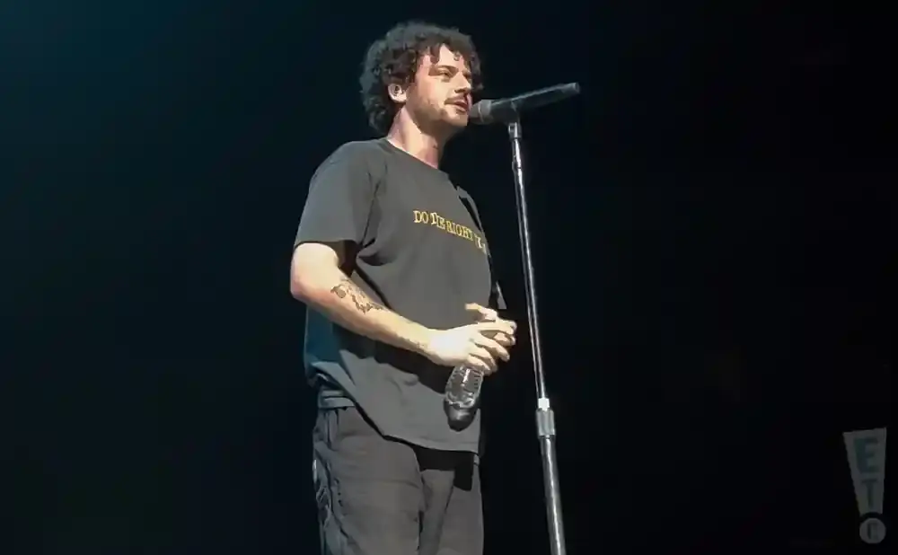 A man with curly hair sings into a microphone on stage, holding a water bottle.