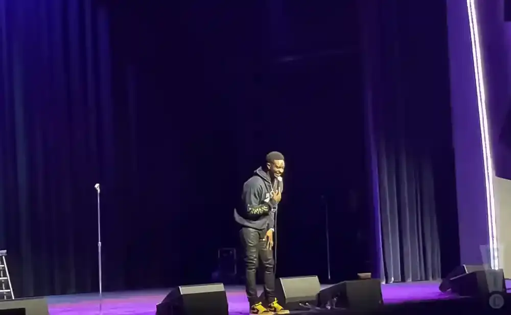A man in a hoodie and leather pants laughs on a stage with purple curtains.