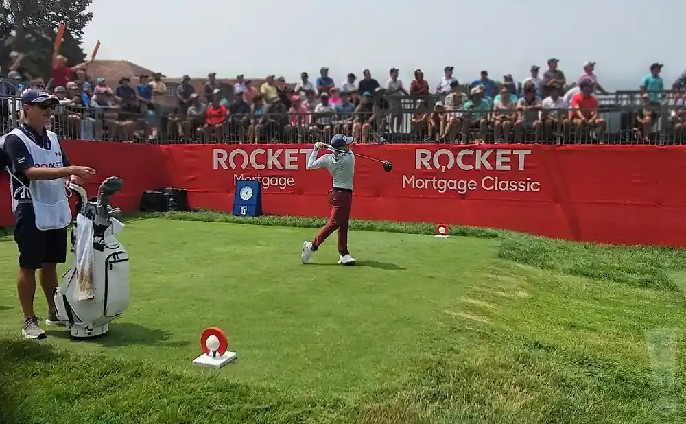 Golfer at Rocket Mortgage Classic swings a club while a caddie stands by a golf bag, with spectators in the background.