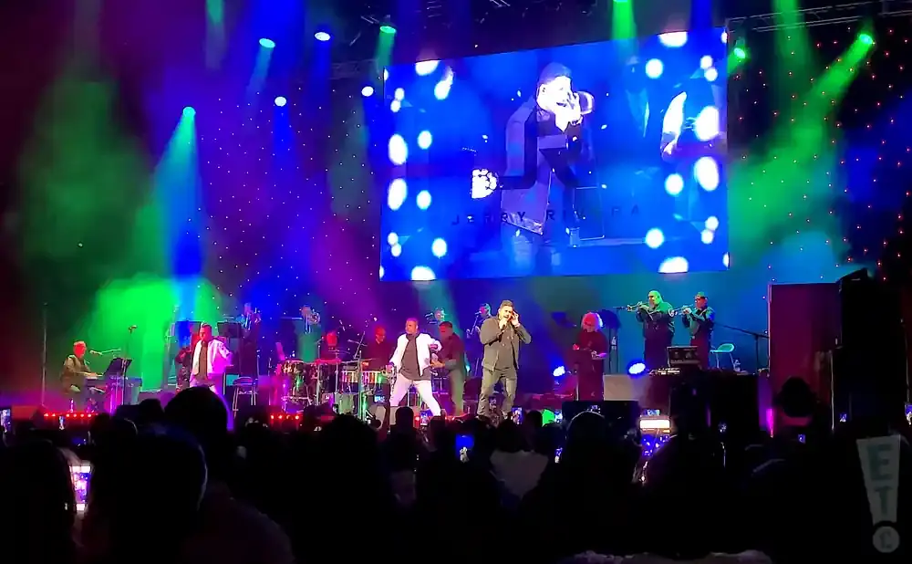Singer Jerry Rivera performs on stage with his band, illuminated by colorful spotlights and a large screen displaying his image.