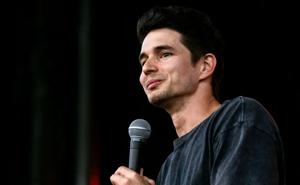 A man smiles while holding a microphone, wearing a dark t-shirt against a dark background.