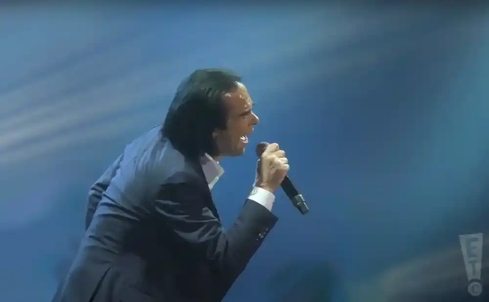 Nick Cave sings into a microphone on a blue-lit stage.