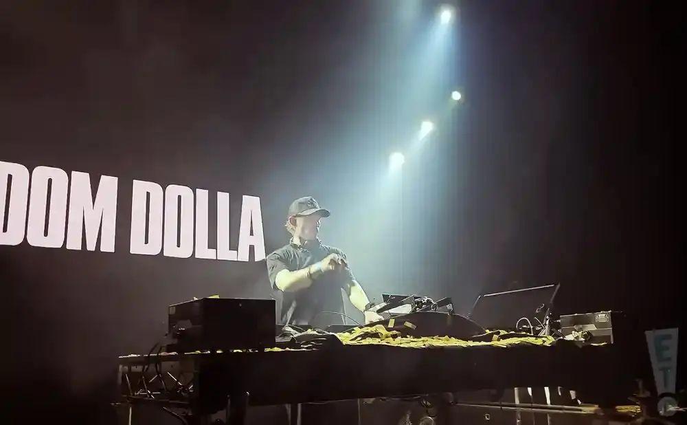 A DJ wearing a black shirt and hat plays music on stage with the name 