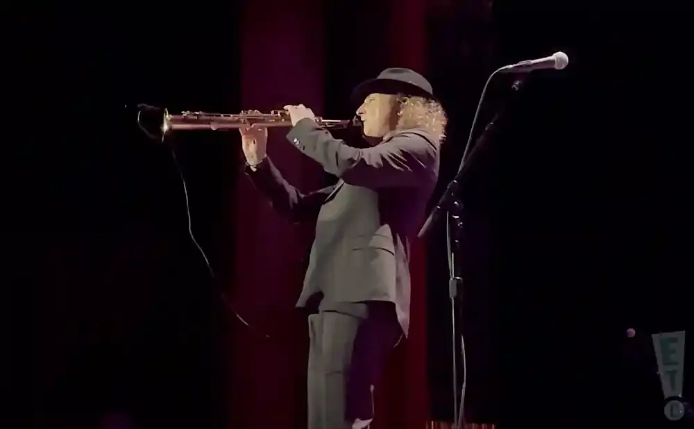 A musician in a hat and suit plays a saxophone on a dimly lit stage.