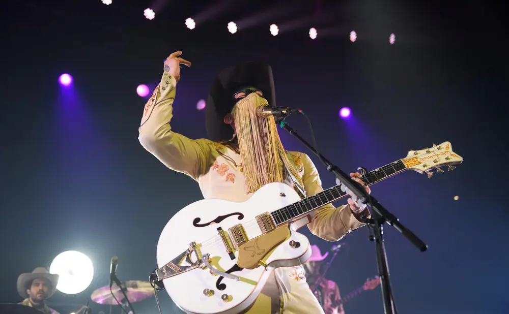 A performer in a cowboy hat and fringe mask plays a white guitar on a stage with purple lights.