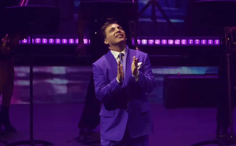 A man in a purple suit and polka-dot tie claps enthusiastically on a dimly lit stage with purple lights.