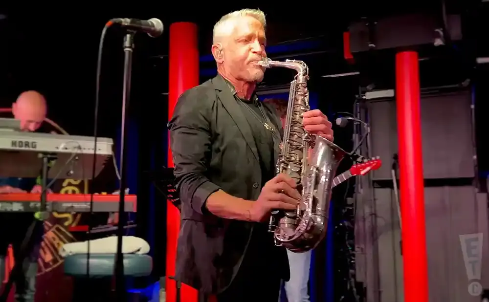 Man plays a saxophone on stage with a keyboardist in the background.
