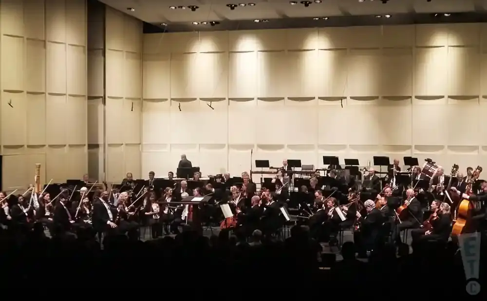 An orchestra performs on a stage with a large wall of light panels behind them.