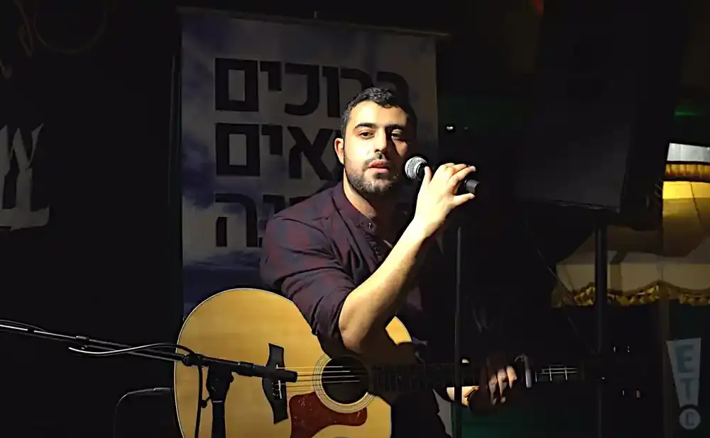 A man with a beard sings into a microphone, playing an acoustic guitar on stage.