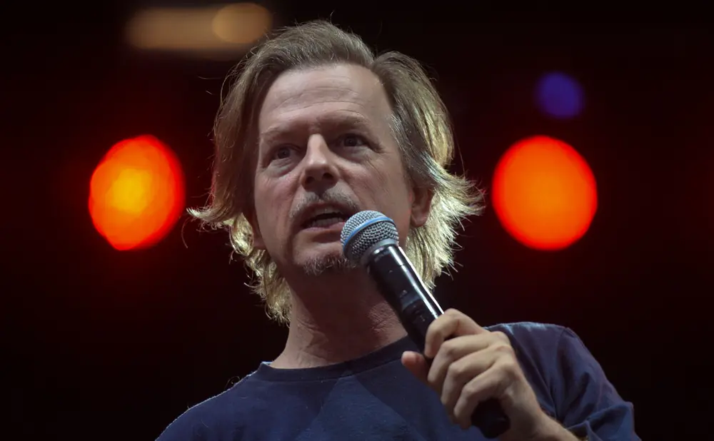 A man with blonde hair and a mustache speaks into a microphone, with red and blue lights behind him.