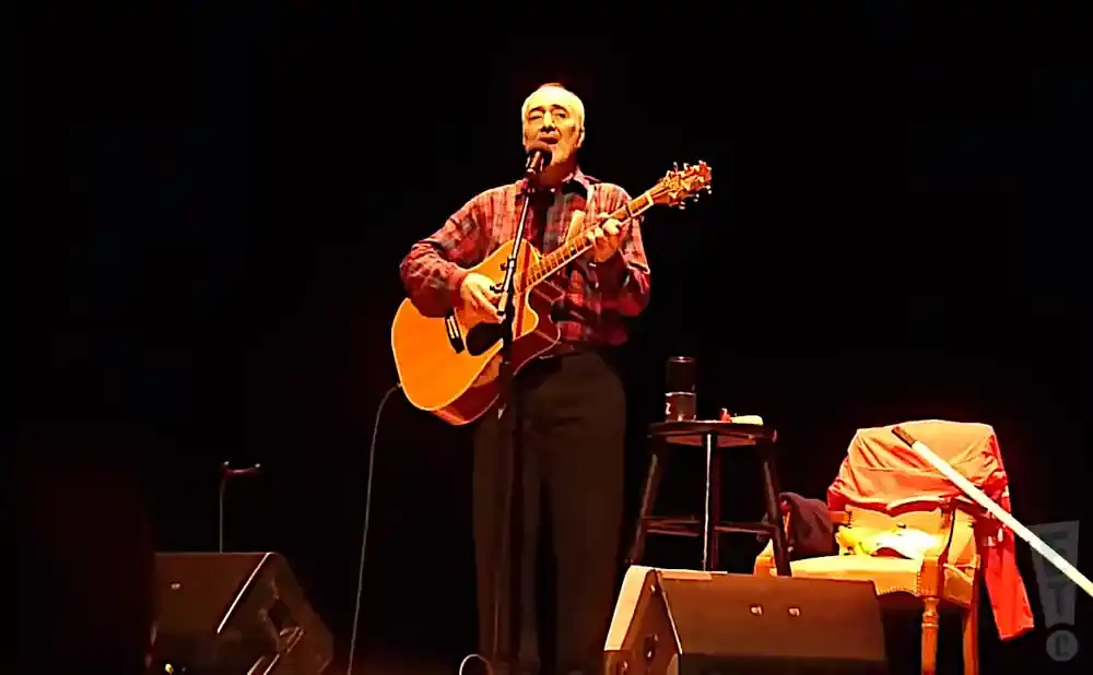 Singer-songwriter plays an acoustic guitar and sings into a microphone on stage.