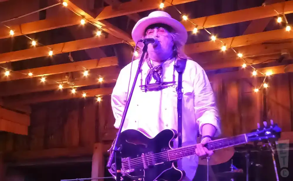 A musician in a white hat and shirt plays a black guitar under string lights.