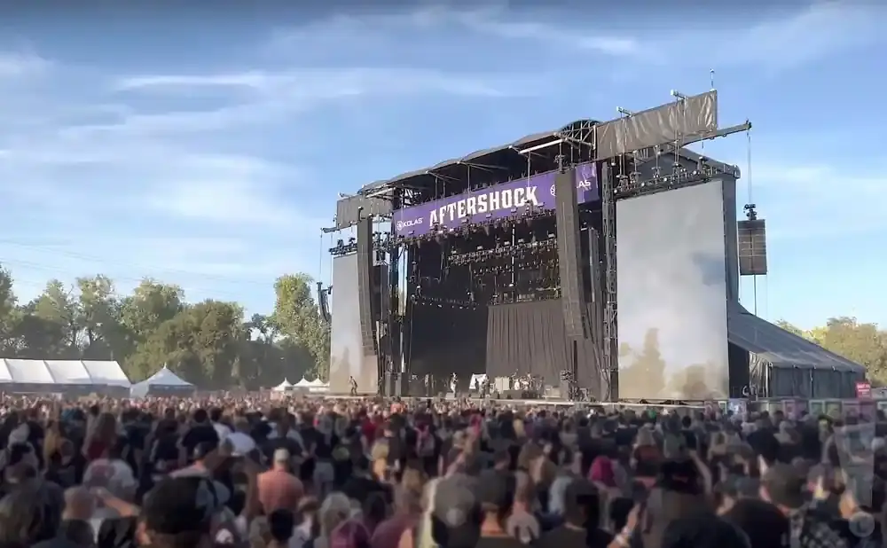 A large outdoor concert crowd watches a band perform on a stage that reads 