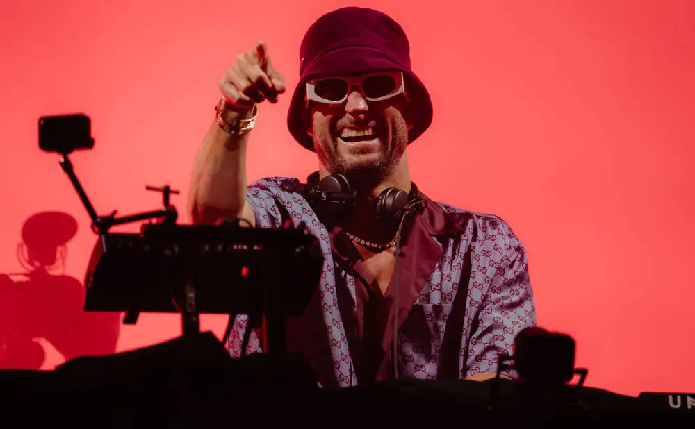 A DJ in a bucket hat and white sunglasses points at the camera, wearing a patterned shirt and headphones.