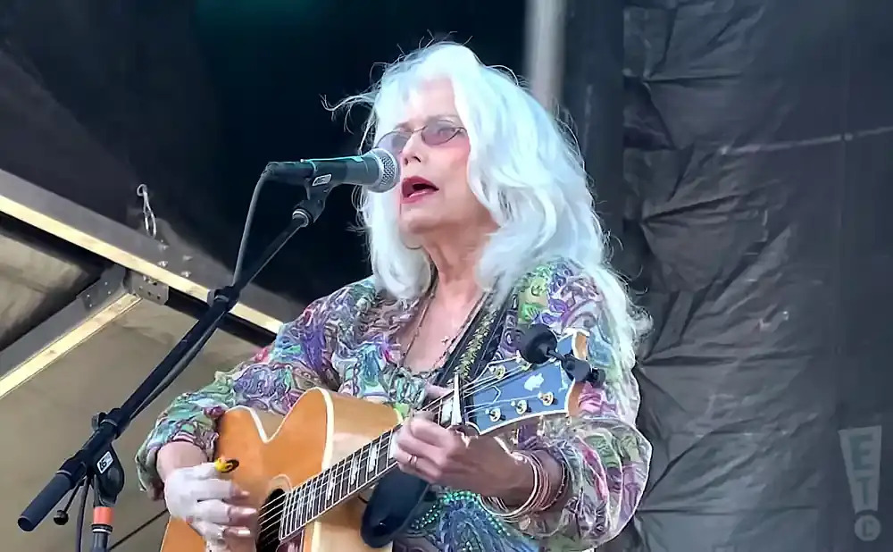 A white-haired woman with glasses sings while playing an acoustic guitar on stage.