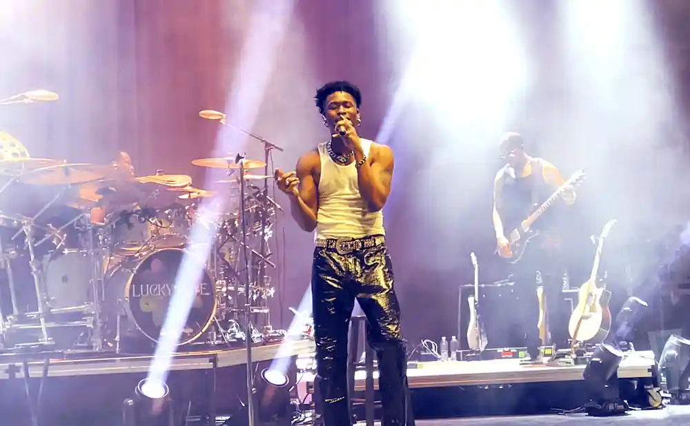 A singer in a white tank top and shiny black pants performs on stage with a band.