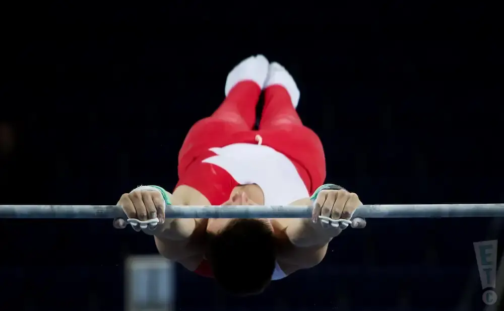 Gymnast in red and white suit performing on the horizontal bar, upside down.
