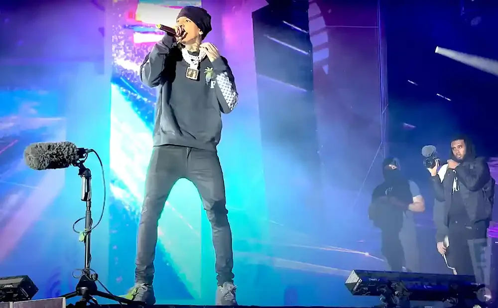A rapper performs on stage with a microphone, wearing a beanie, hoodie, and slim-fit pants.