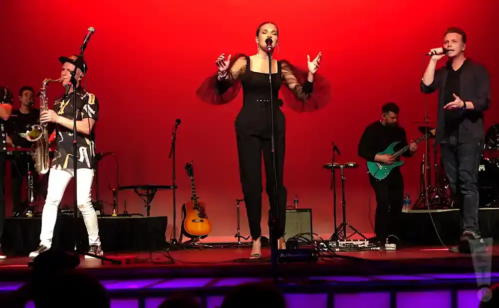 Band performing on a stage with red backdrop. A woman in black sings center, with a saxophonist left and a guitarist right.