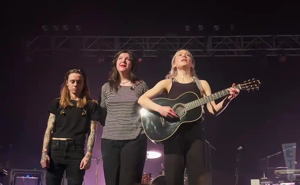Three musicians on stage: one with an acoustic guitar, one in a striped shirt, and one with long hair and tattoos.
