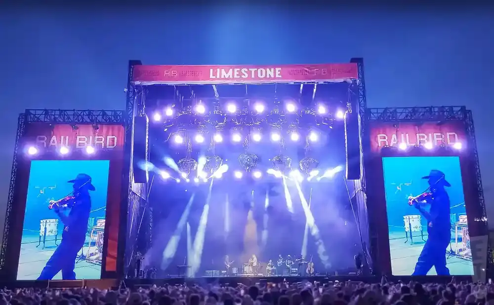 A live music festival stage with large screens showing a violinist, under a blue evening sky.