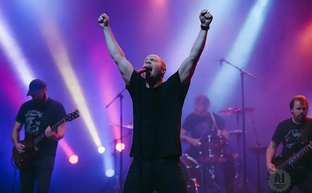 A male singer with arms raised sings into a microphone on a stage with colorful lighting.