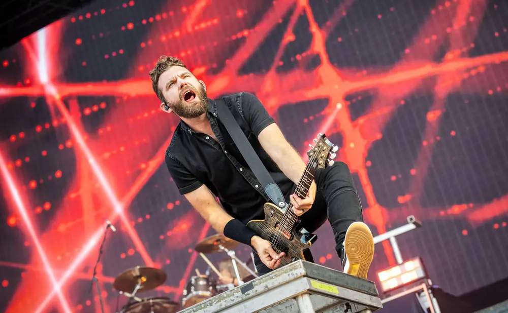 A male guitarist plays an electric guitar on a stage with a red, abstract background at a concert.