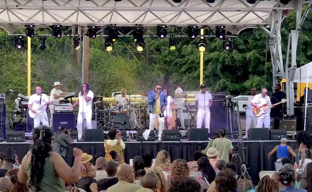 A band performs on an outdoor stage at a concert, with an audience in the foreground.