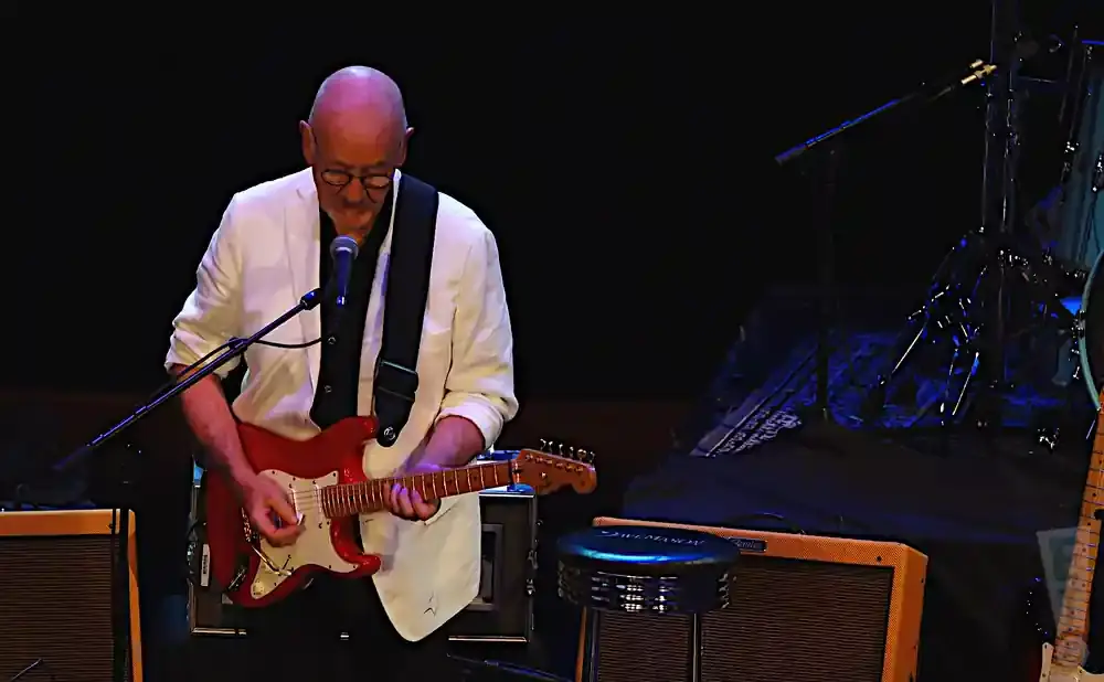A bald man in a white jacket plays a red electric guitar on stage.