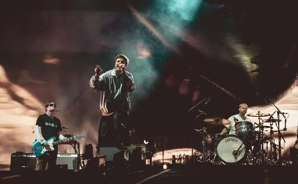 A band performs on a dimly lit stage with a large screen behind them. The lead singer, in a tracksuit, sings into a microphone.