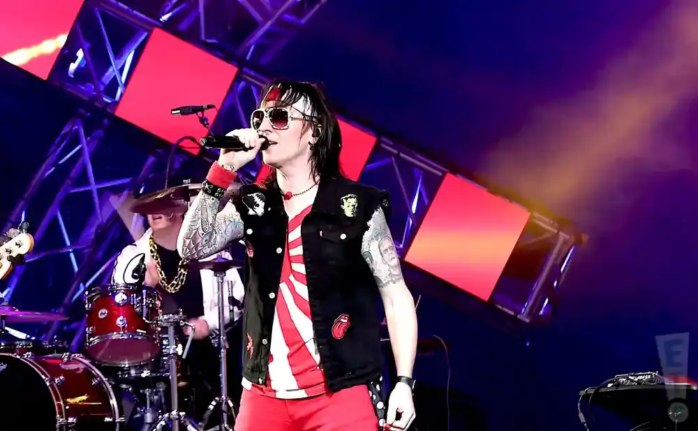 A rock singer with sunglasses and tattoos performs on stage, wearing a denim vest over a red and white striped shirt and red shorts.