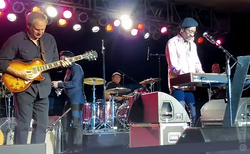 A band performs on stage with a guitarist, keyboardist, and drummer under bright lights.