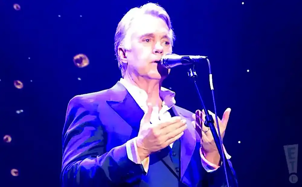 Man in a suit singing into a microphone on a blue stage with floating lights.
