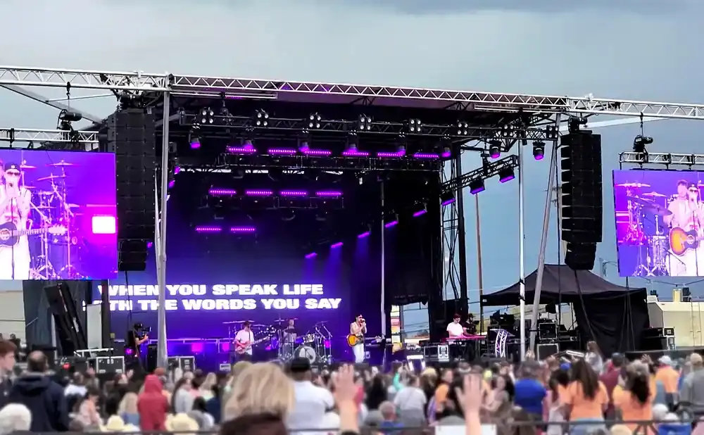 Band performs on a stage with large screens and purple lighting, in front of a crowd.