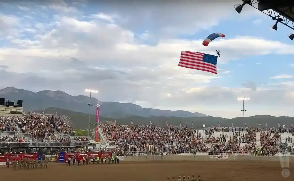 A skydiver with a U.S. flag descends toward a crowded stadium with mountains in the background.